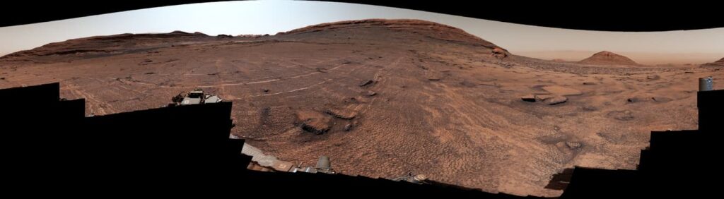 Panorama du sol rocheux de Mars couvert de petites fractures polygonales, avec des collines à l’horizon et la butte Miraflores sur la droite.