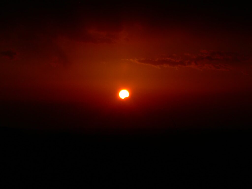 Soleil orange partiellement masqué par la Lune, très bas au-dessus d’un horizon sombre, dans un ciel rougeoyant au coucher du Soleil.