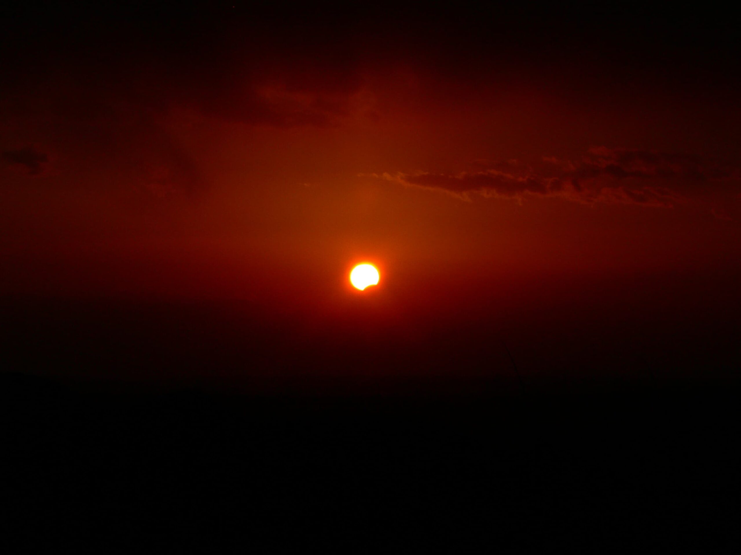 Soleil orange partiellement masqué par la Lune, très bas au-dessus d’un horizon sombre, dans un ciel rougeoyant au coucher du Soleil.