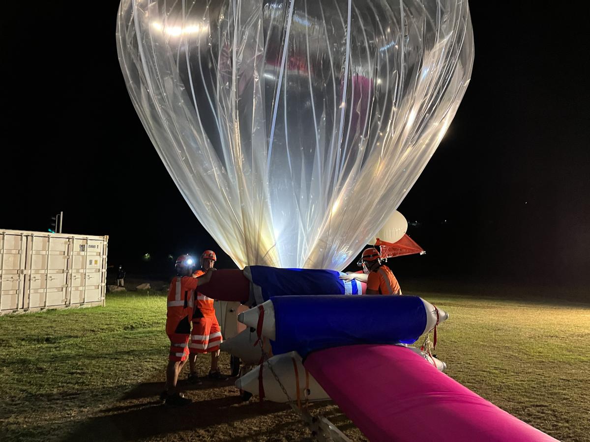 Équipe en tenues de sécurité gonflant l’enveloppe transparente d’un ballon Strateole‑2 la nuit à Mahé, avec la nacelle et le matériel au sol.