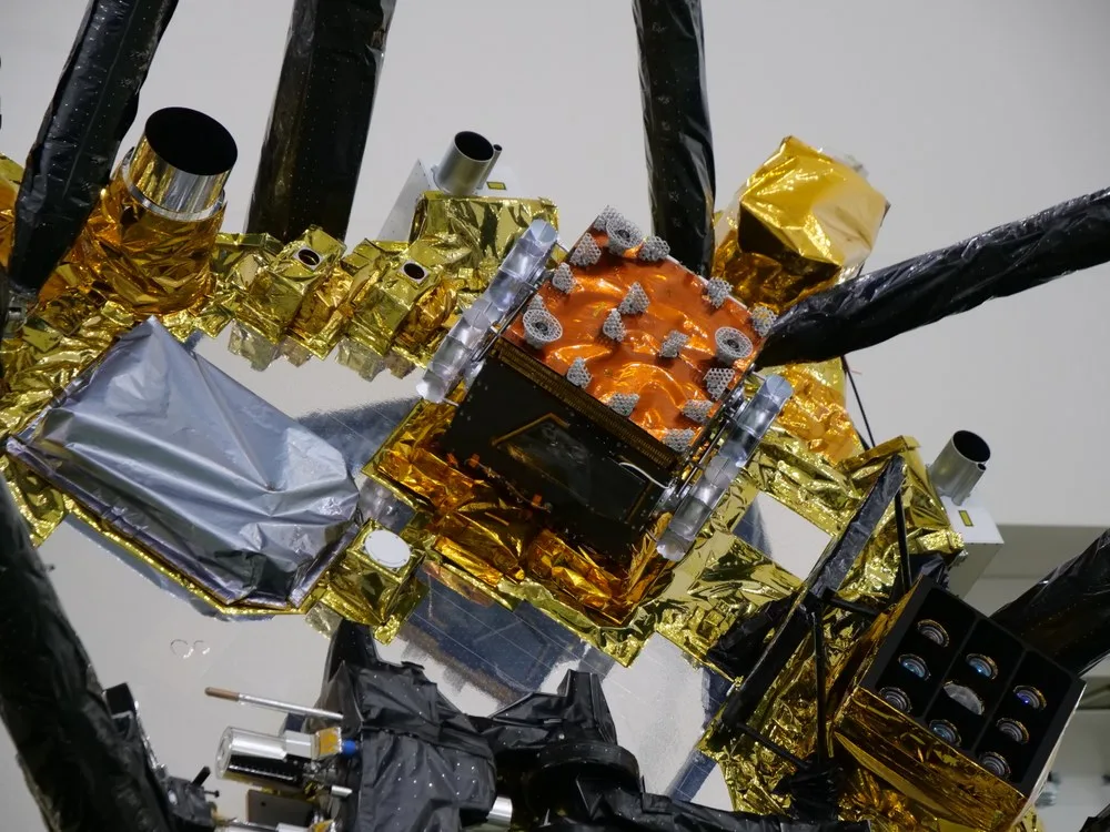 Petit rover Idefix installé en position de voyage entre les jambes d’atterrissage de la sonde MMX, boîtier compact orange et composants recouverts de films métallisés dorés et argentés.