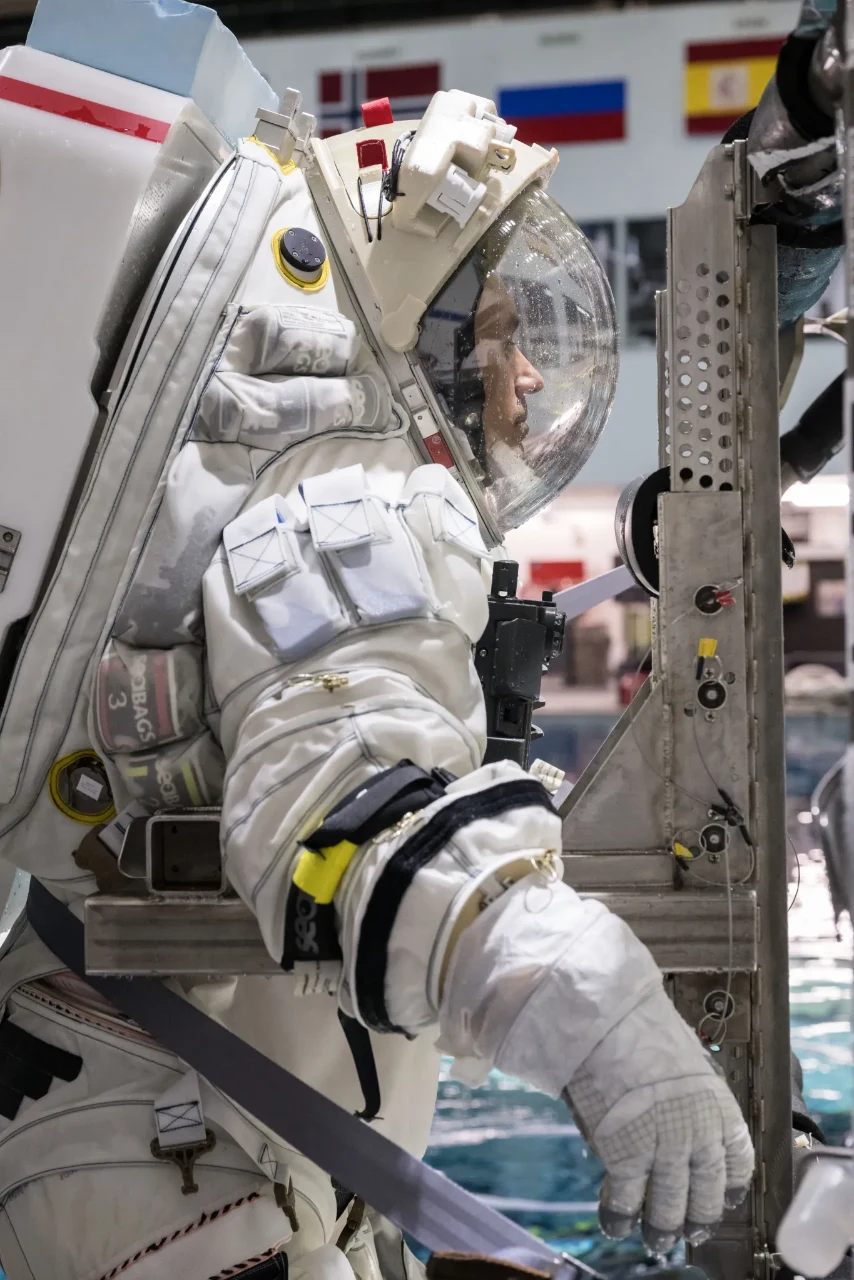 Richard Rhodes en combinaison lunaire d’Axiom Space, casque fermé avec des gouttelettes d’eau, s’entraînant au bord d’une structure au-dessus d’une piscine.