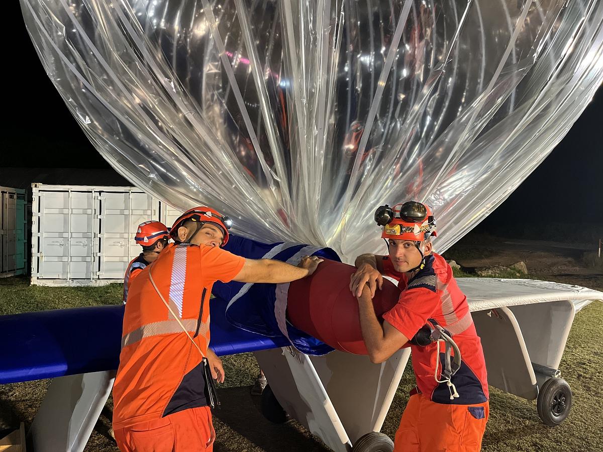 Des ballonniers du CNES en tenue orange et casques éclairés contrôlent l’enrobage d’une nacelle sous l’enveloppe transparente d’un ballon pressurisé Strateole‑2 la nuit, sur l’aire de lancement aux Seychelles.