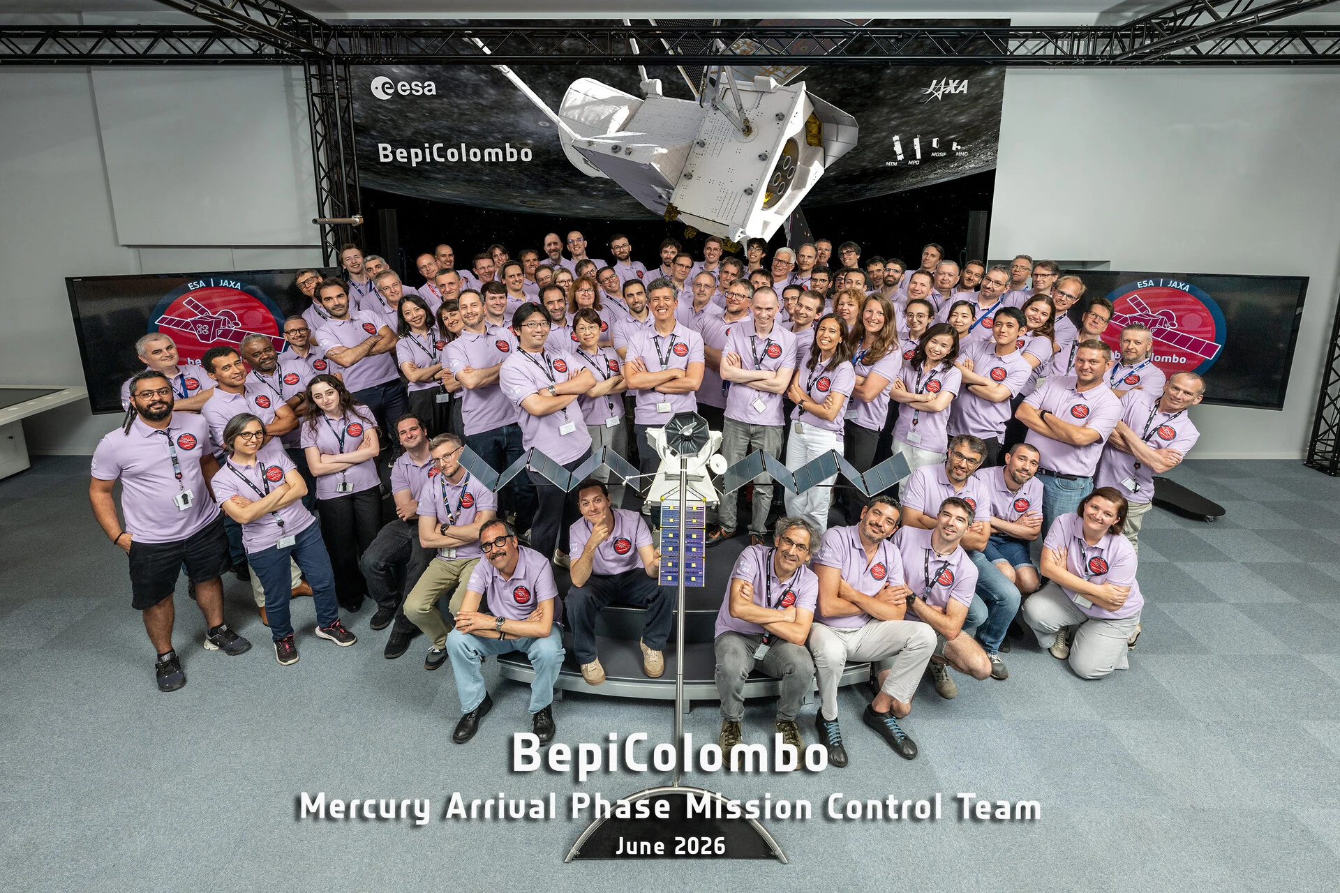 Membres de l’équipe de contrôle de BepiColombo réunis au Centre européen des opérations spatiales, posant devant une maquette et un grand visuel de la mission.