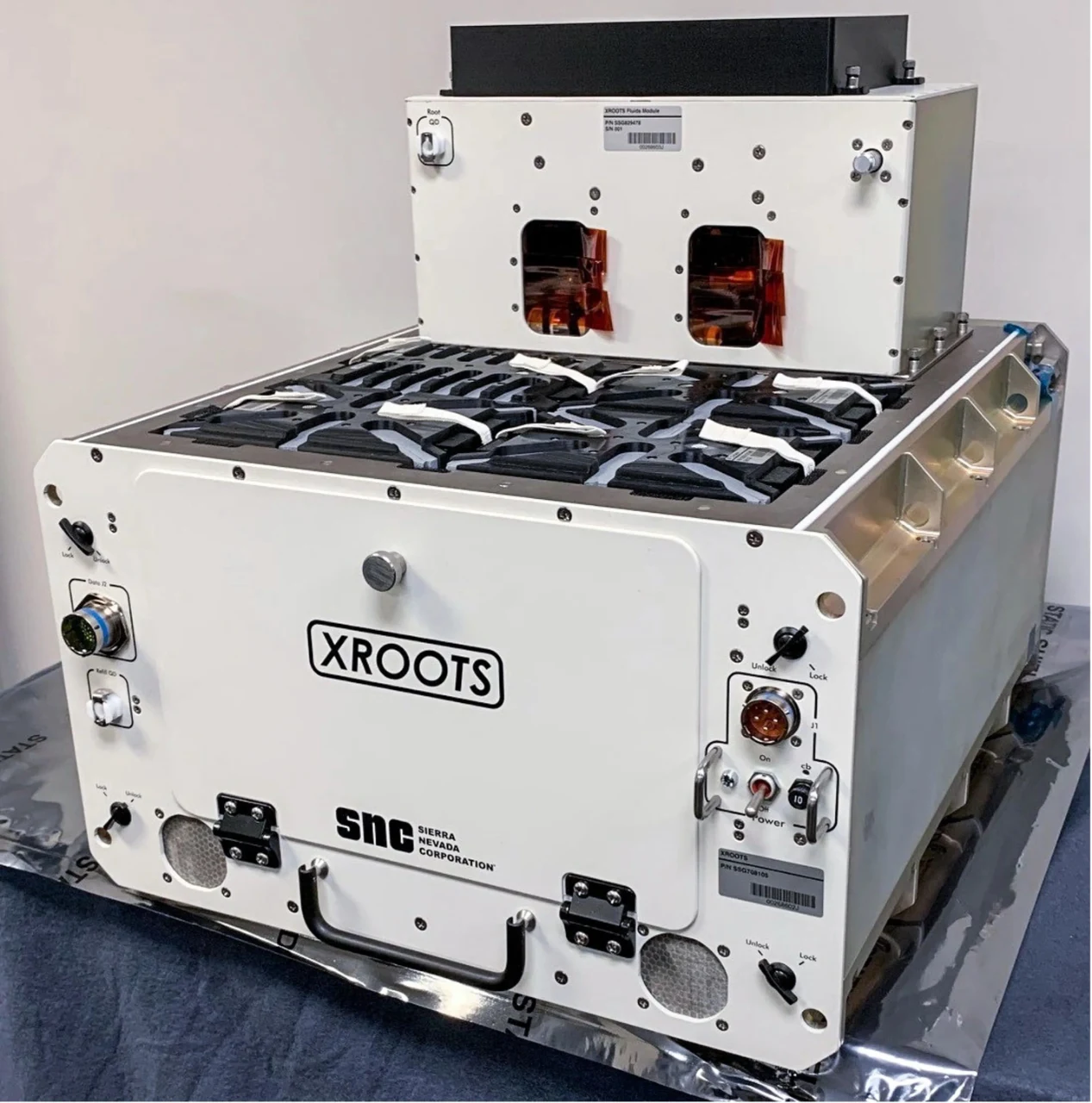 Grand boîtier métallique blanc portant l’inscription « XROOTS », avec un module plus petit fixé sur la partie supérieure et plusieurs emplacements de chambres de croissance visibles sur la surface supérieure.