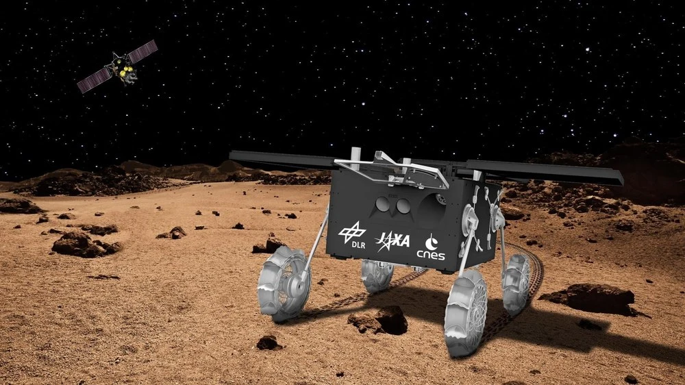 Vue d’artiste du rover Idefix debout sur le sol de Phobos, la sonde MMX en arrière-plan.