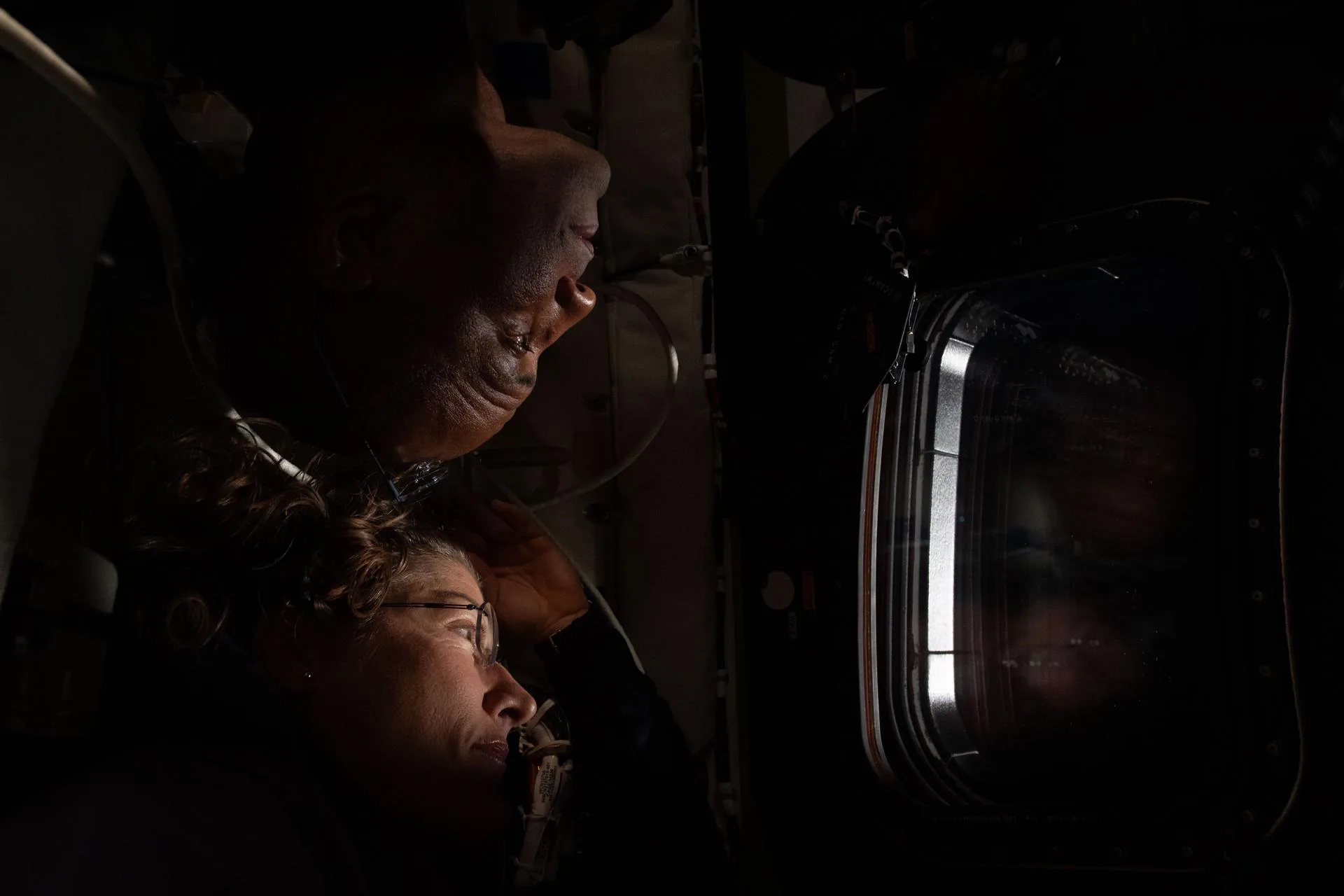 Christina Koch (en bas) et Victor Glover (en haut) regardent côte à côte par un hublot de la capsule Orion, visages éclairés par la lumière extérieure pendant le survol lunaire.
