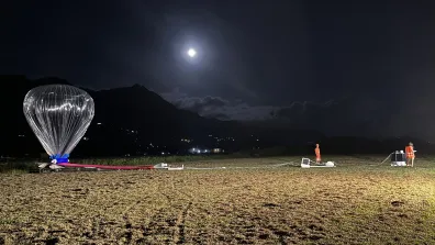 Ballon pressurisé prêt au lâcher la nuit sur une aire d’herbe, relié par des lignes et équipements au sol, techniciens et projecteurs autour, paysage montagneux en arrière‑plan et pleine lune dans le ciel.
